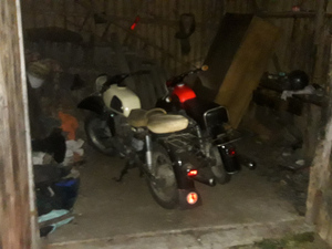 dwa motocykle MZ stojące obok siebie w drewnianym budynku gospodarczym na jednej z posesji. Motocykl z lewej strony jest koloru białego a po prawej koloru czarno-czerwonego